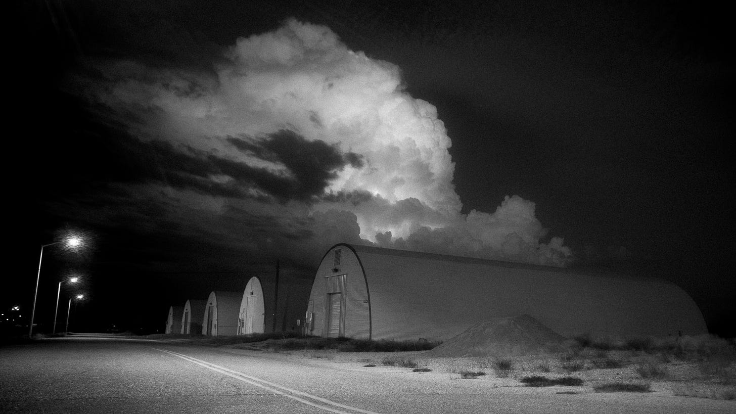 A high-contrast black-and-white night photograph showing a row of corrugated metal Quonset huts alongside an empty asphalt road. In the pitch-black sky behind the structures, a massive, brightly illuminated cumulonimbus thunderhead billows upward. A few glowing streetlights cast light along the road on the left. A high-contrast black-and-white night photograph showing a row of corrugated metal Quonset huts alongside an empty asphalt road. In the pitch-black sky behind the structures, a massive, brightly illuminated cumulonimbus thunderhead billows upward. A few glowing streetlights cast light along the road on the left.