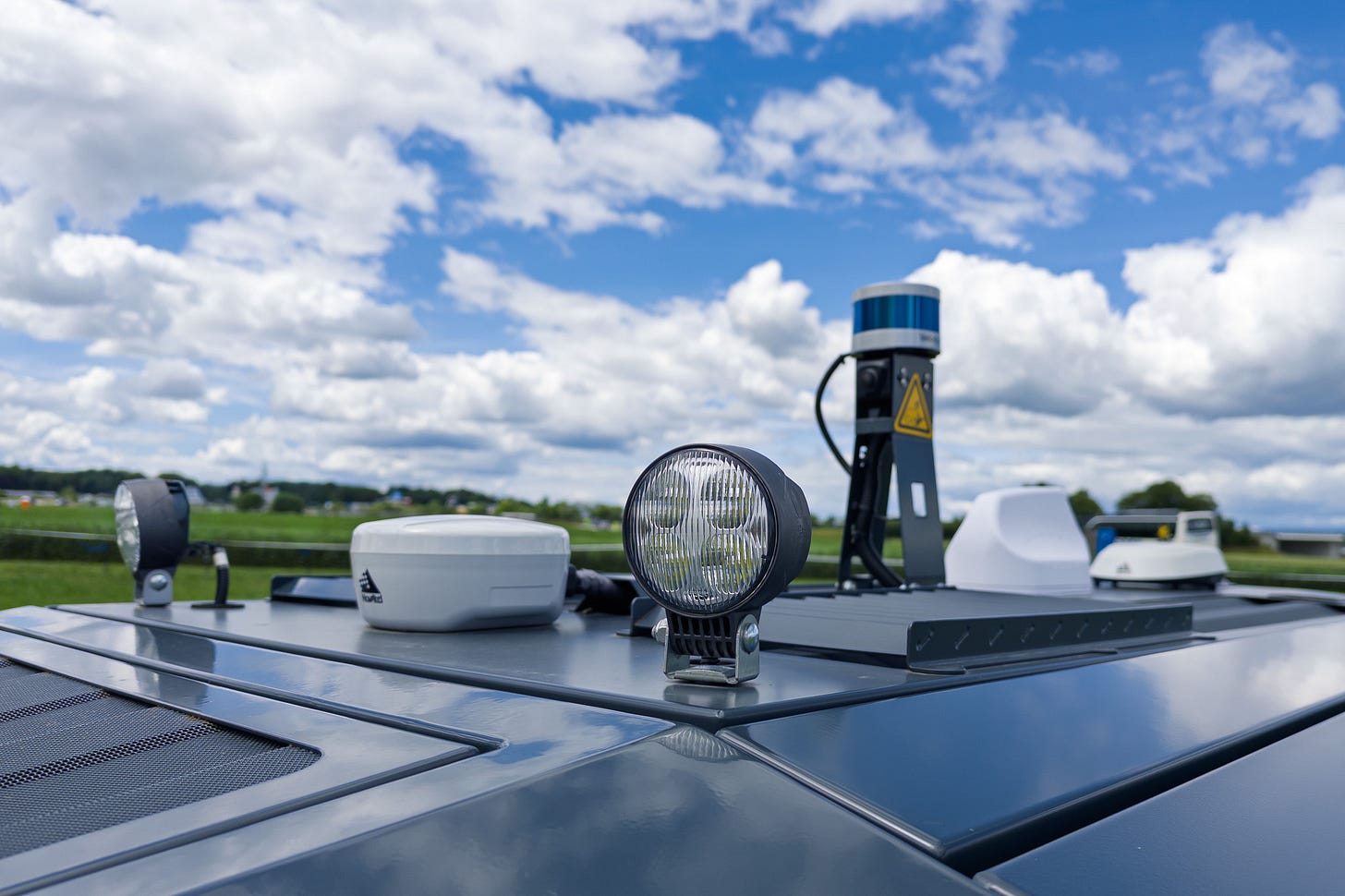 Das Bild zeigt die technische Ausrüstung des autonomen Feldroboters AgBot von AgXeed. Auf dem Dach sind Scheinwerfer und ein 3D-LiDAR-Sensor zu sehen, welcher der Umgebungserfassung dient. Die glänzende Metalloberfläche spiegelt Licht und Wolken; dahinter öffnet sich eine weite Agrarlandschaft.