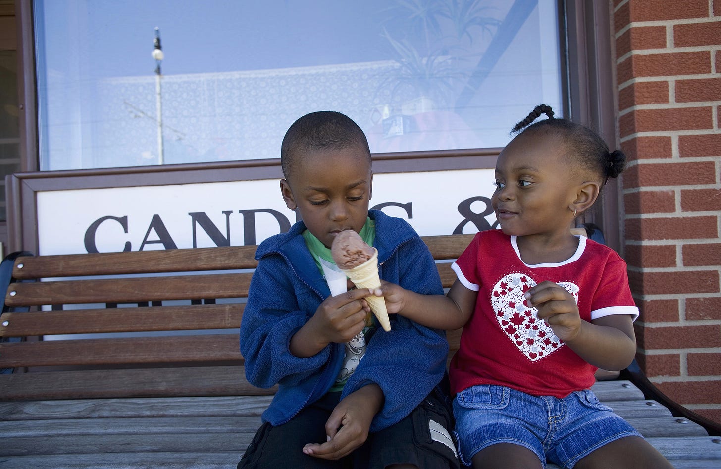 A little girl on a bench offering a lick of her ice cream to the boy sitting next to her