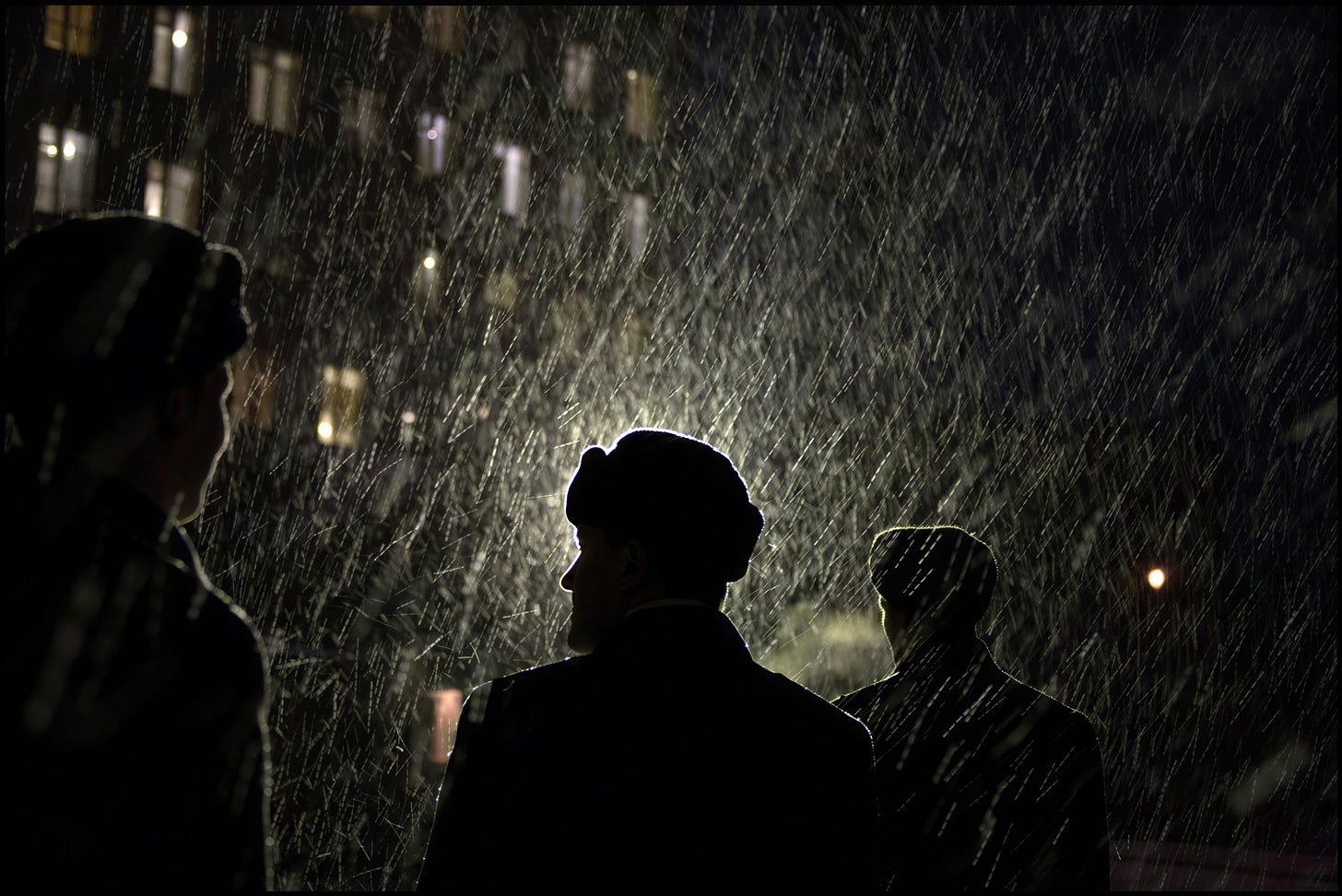 Gueorgui Pinkhassov - Three people stand in the dark, their silhouettes illuminated by a bright light behind them as heavy snow falls, with blurred building windows lit in the background.