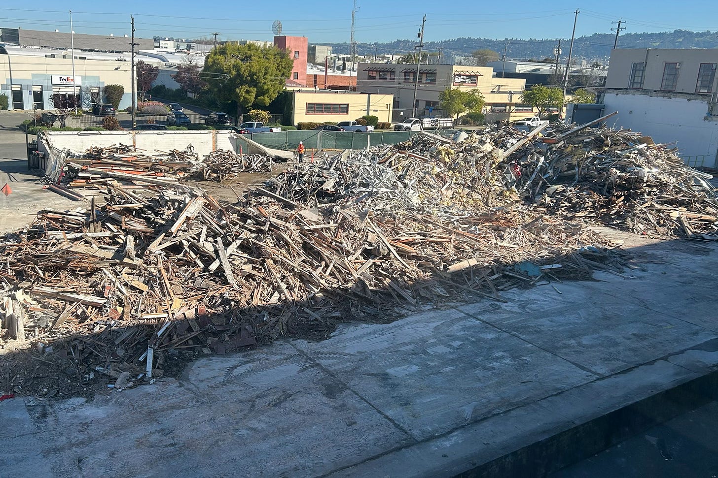 A large demolition site is filled with multiple piles of debris consisting of splintered wood, twisted metal, insulation, and other building materials from a recently demolished structure. In the background, two construction workers in orange safety vests are visible near the remaining foundation walls. Surrounding the site are industrial and commercial buildings, including a FedEx facility and a beige and red fire station, set against a backdrop of East Bay hills and clear blue sky. The foreground includes a concrete slab with visible tire tracks from heavy machinery. The scene captures urban demolition, construction waste, and site clearance in progress.