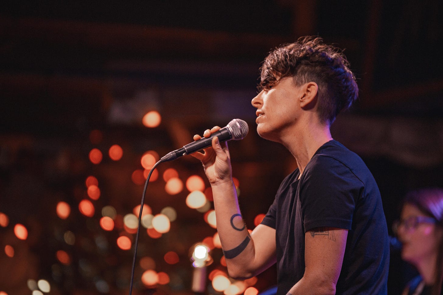 This photo. Image description: Andrea holds a mic while wearing a black tshirt. Colorful lights in the background. This photo. Image description: Andrea holds a mic while wearing a black tshirt. Colorful lights in the background.