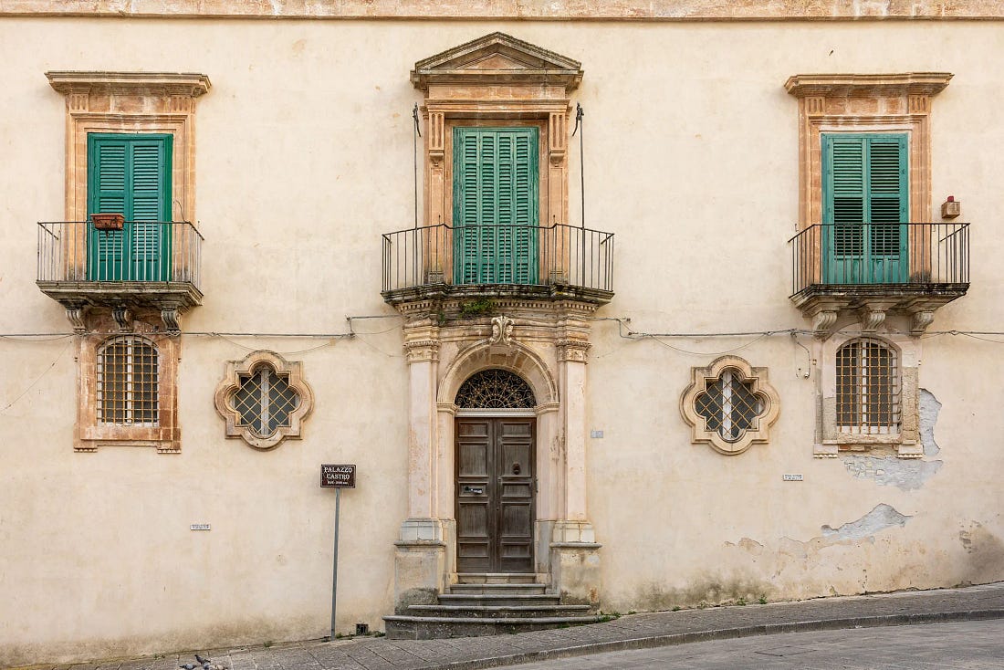 The cream facade of a baroque palazzo in Ragusa Ibla, with teal shutters behind iron balconies, carved stone window frames, quatrefoil windows and an arched wooden door. The cream facade of a baroque palazzo in Ragusa Ibla, with teal shutters behind iron balconies, carved stone window frames, quatrefoil windows and an arched wooden door.