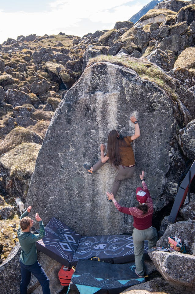 A climber with long dark hair, seen from behind in a brown shirt and grey pants, moves up the steep face of a tall grey boulder. Below, two spotters stand with arms raised: one in a green hoodie and jeans, one in a dark red patterned sweater and cap. Thick bouldering pads with geometric patterns cover the landing. A small white dog watches from the rocks at left. Boulderfield and sunlit tundra stretch behind. A climber with long dark hair, seen from behind in a brown shirt and grey pants, moves up the steep face of a tall grey boulder. Below, two spotters stand with arms raised: one in a green hoodie and jeans, one in a dark red patterned sweater and cap. Thick bouldering pads with geometric patterns cover the landing. A small white dog watches from the rocks at left. Boulderfield and sunlit tundra stretch behind.