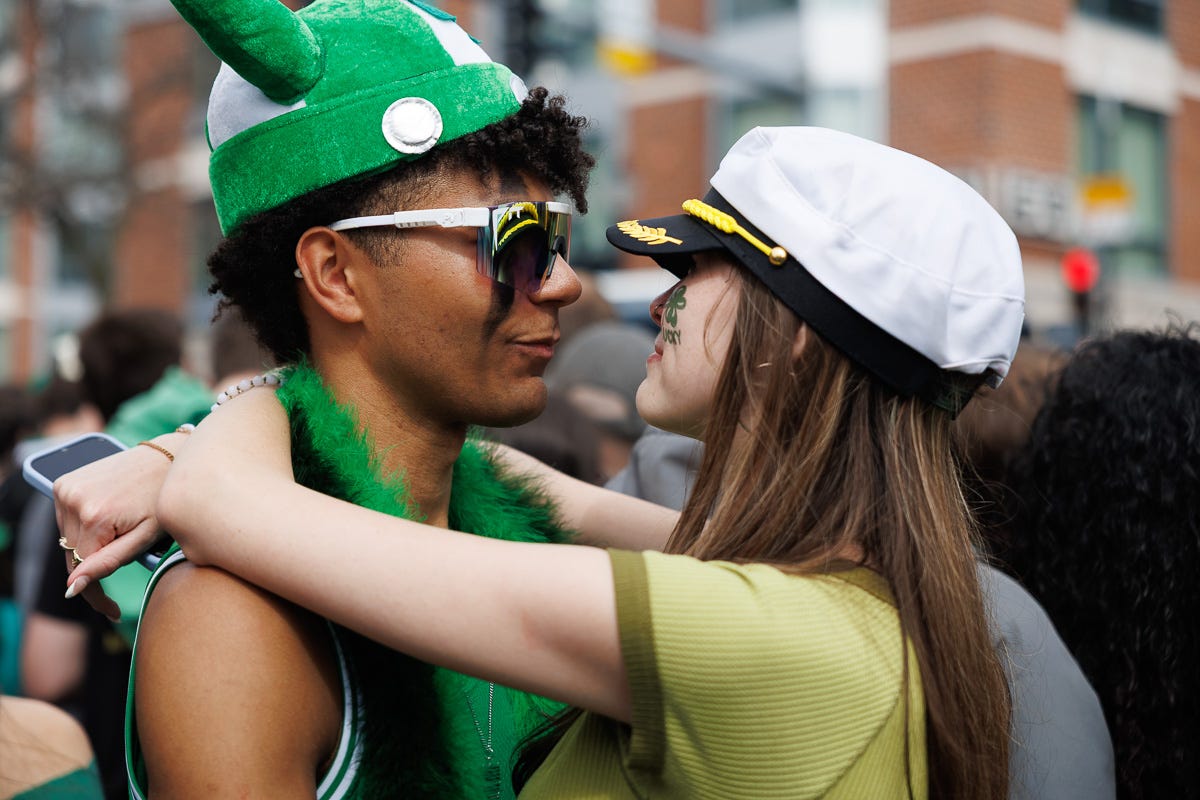 A young couple in festive green outfits stares into each other’s eyes, their faces close together, surrounded by a street crowd. A young couple in festive green outfits stares into each other’s eyes, their faces close together, surrounded by a street crowd.