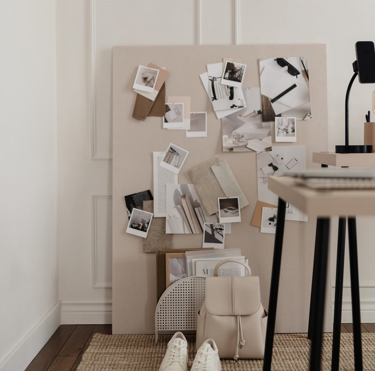A neutral-toned creative workspace featuring a vision board with pinned photos, fabric swatches, and paper samples. A pair of white sneakers and a beige backpack rest on a woven rug beside a light wood desk.