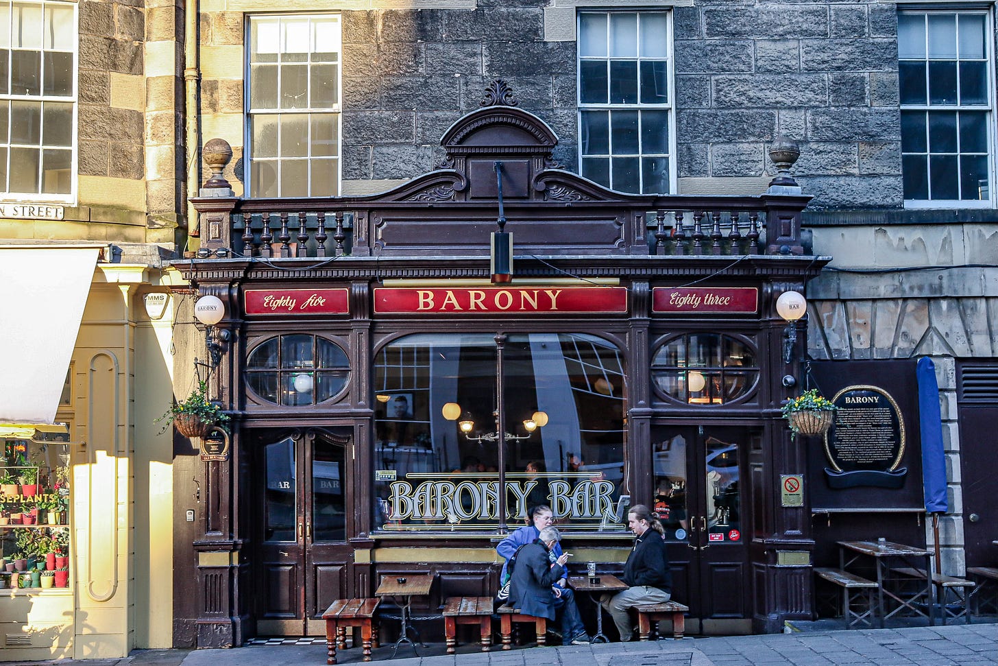 An ornate teak frontage of a pub at street level with a red and gold sign. Three people sit at the benches outside.