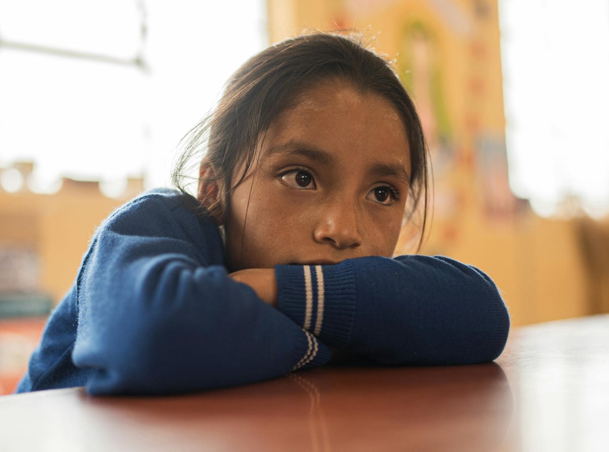 Child sitting at table, resting her head on crossed arms, looking bored