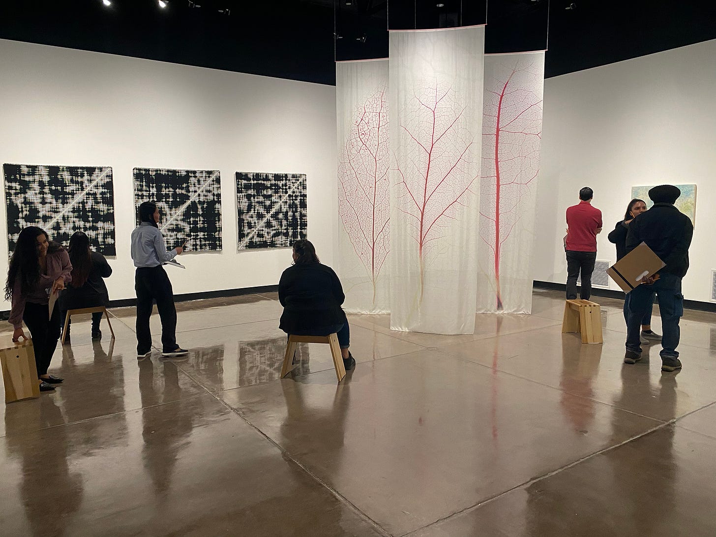 Participants in a gallery with white walls and a polished concrete floor, several seated on low wooden stools, some holding clipboards. They face three large black-and-white textile works on the left wall and three tall translucent banners printed with red leaf veins hanging at center. Others stand and look toward a painting on the right wall.