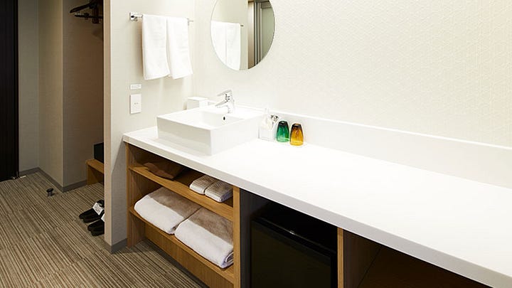 The modern, bright reception area of Hotel Intergate Kanazawa featuring a colorful geometric tiled wall and a long minimalist wooden front desk. An accessible hotel bathroom vanity area with a wide white countertop, a square vessel sink, and open wooden shelving containing towels and a small refrigerator. A spacious accessible hotel room featuring two twin beds, a large brown sofa, and a small round table with colorful patterned chairs and stools. An accessible bathroom featuring a bathtub with a handheld shower head, a sink, and a toilet with grab bars against light wood-textured walls.