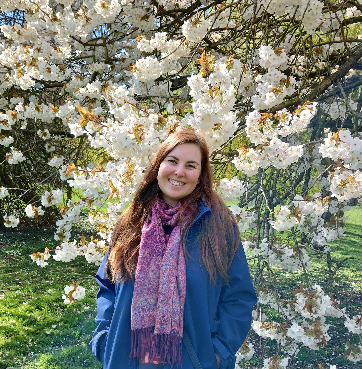 smiling brunette woman in a blue coat and purple scarf standing before white blossom