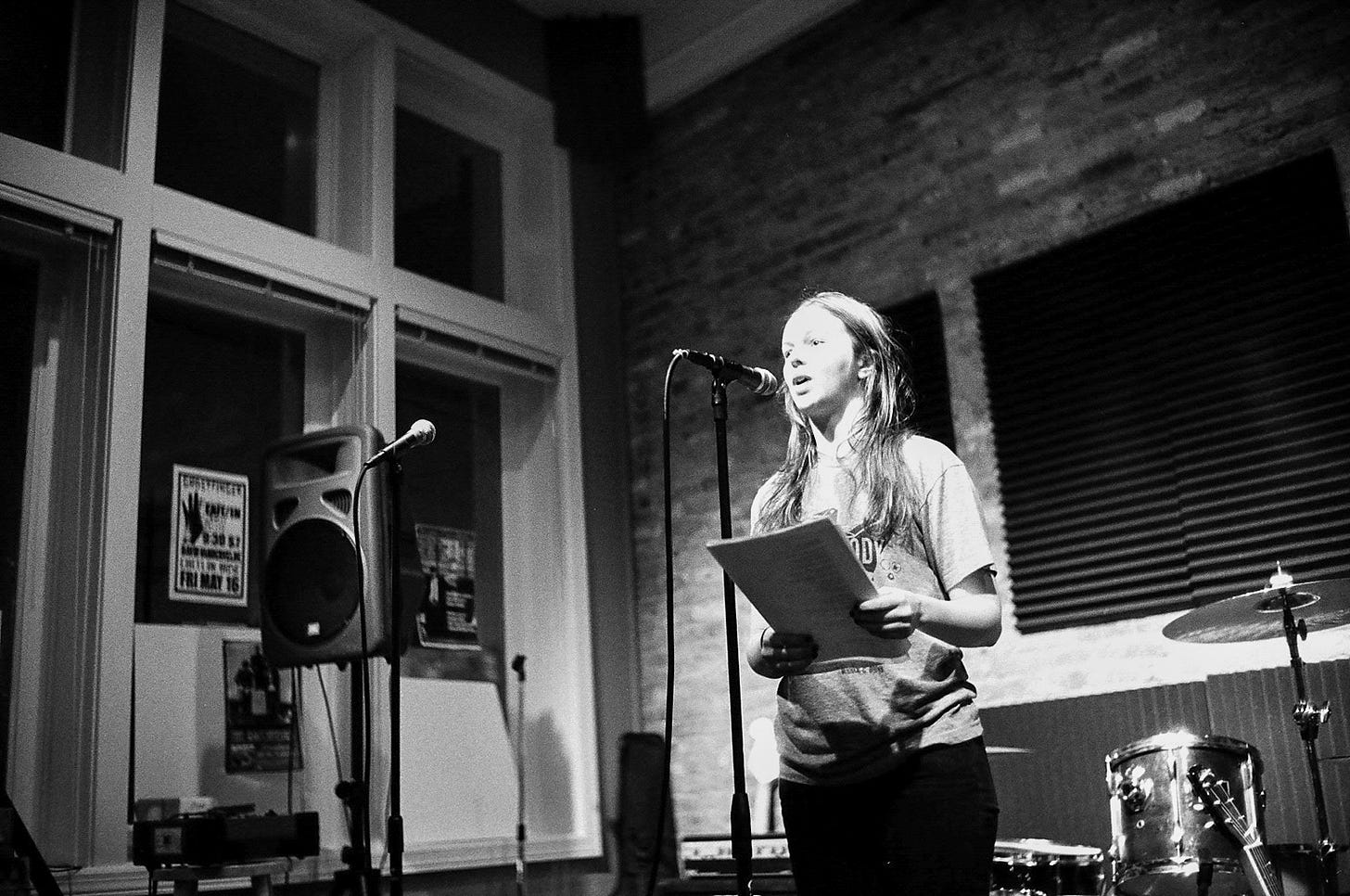 Amber is speaking into a microphone on a stand. She holds papers that she is reading from. Speakers, additional microphones, and a drum kit are seen behind her.