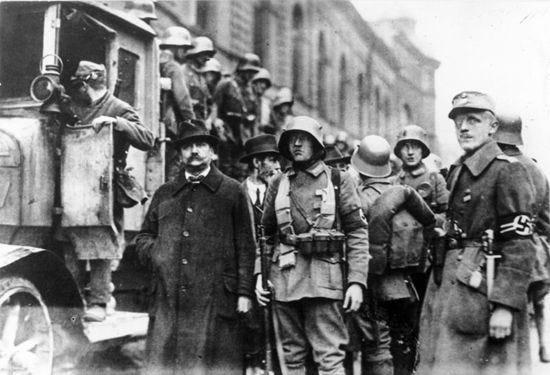 Historical black and white photograph from 9 November 1923 showing Adolf Hitler and armed supporters wearing helmets and Nazi armbands during the Beer Hall Putsch march through Munich streets.