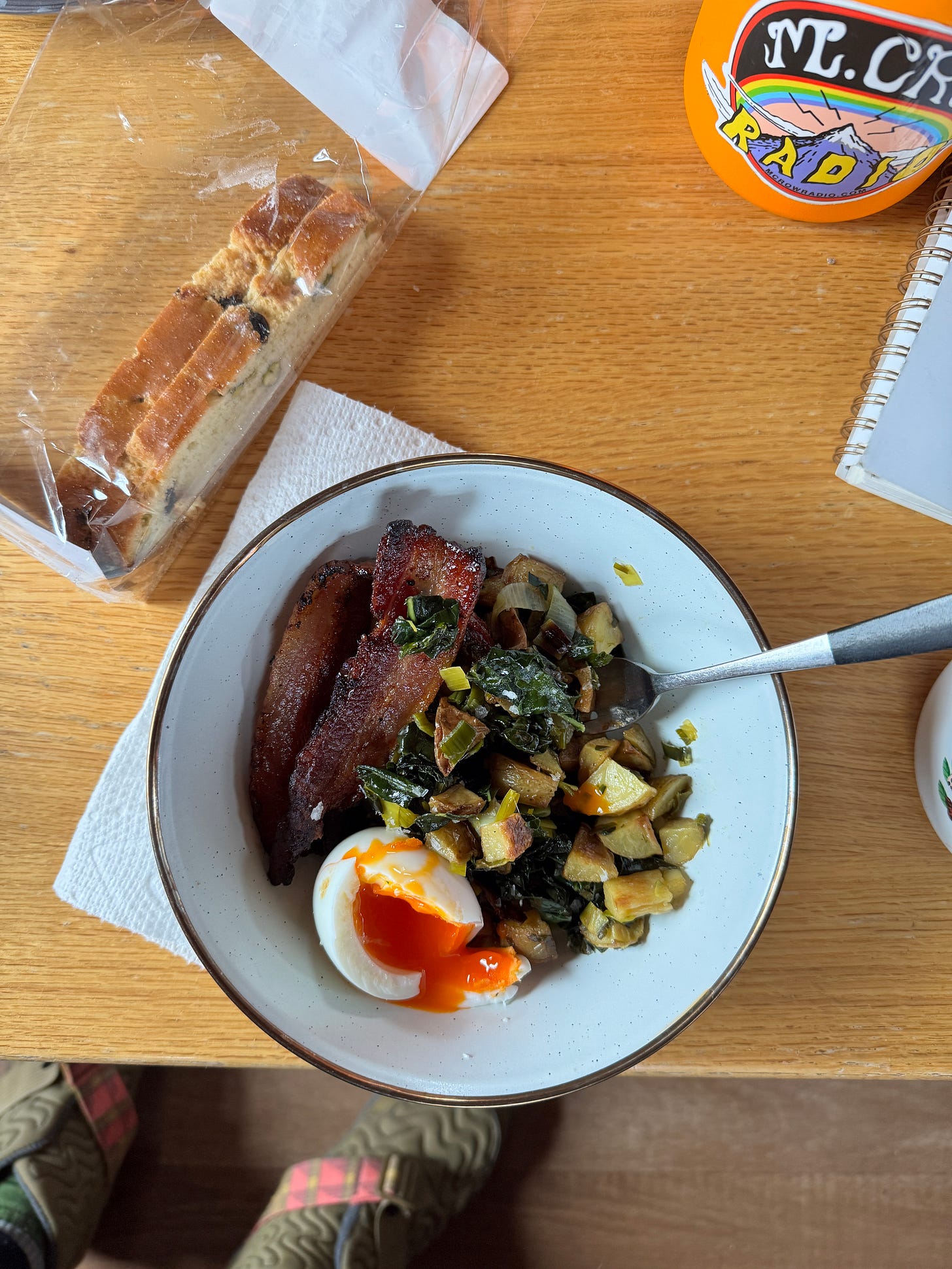 Overhead shot of a bowl filled with potato and kale hash, bacon, and a soft-cooked egg broken open to reveal a bright orange yolk. The bowl is on a wood table next to a cellophane bag with a couple of biscotti inside, a water bottle, and the corner of a spiral-bound notebook. Overhead shot of a bowl filled with potato and kale hash, bacon, and a soft-cooked egg broken open to reveal a bright orange yolk. The bowl is on a wood table next to a cellophane bag with a couple of biscotti inside, a water bottle, and the corner of a spiral-bound notebook.