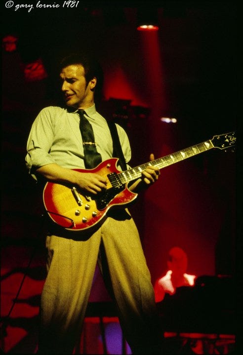 Colour photo of Midge Ure on stage playing guitar.