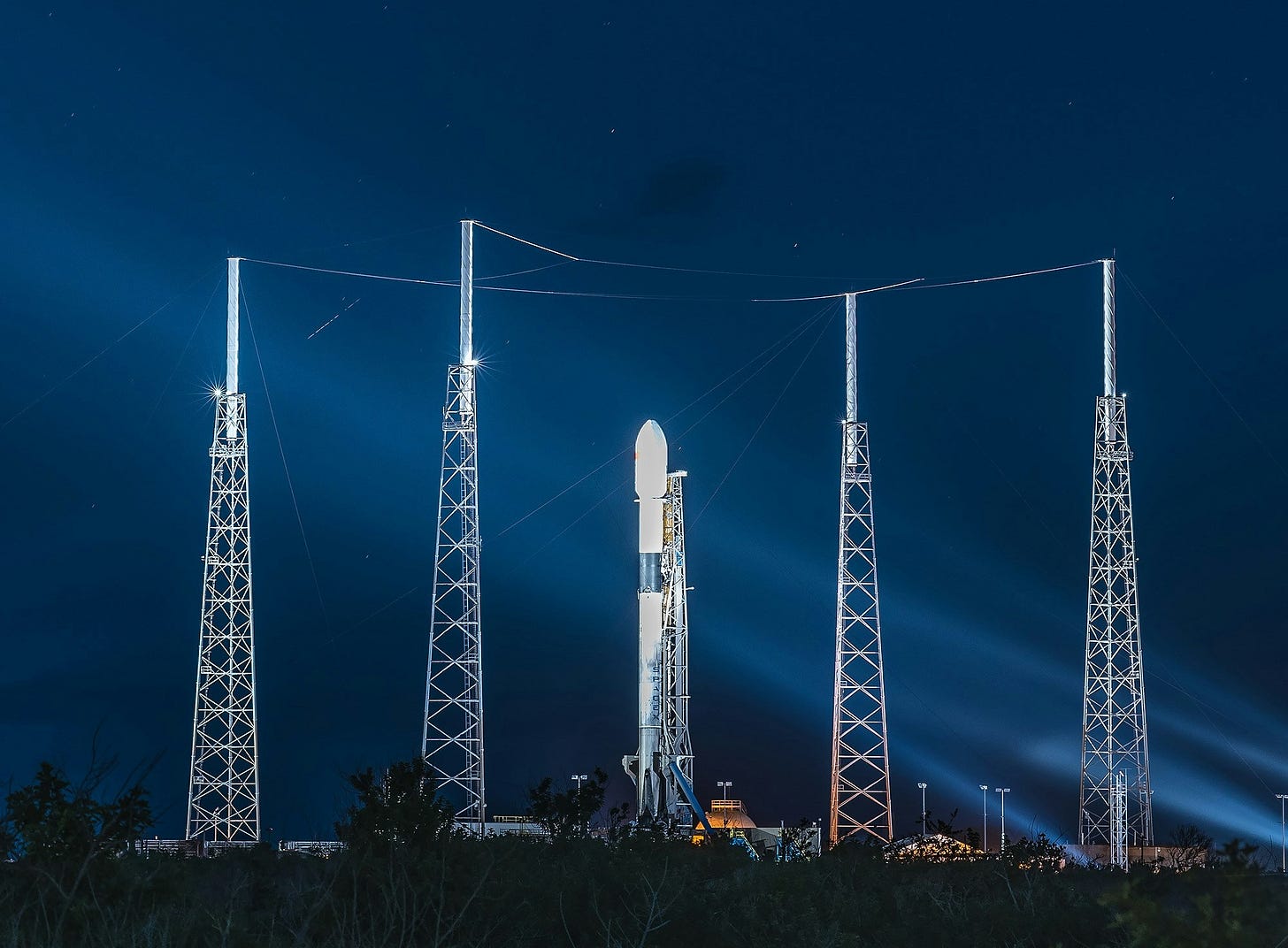 A vertical rocket stands on a launchpad at night, illuminated by bright blue spotlights under a starry sky. The rocket is flanked by four tall, skeletal metal lightning towers connected by wires, with dark tropical foliage in the foreground.