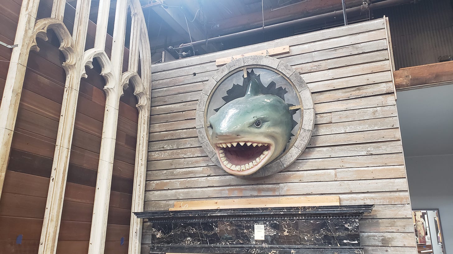 Fake gray shark head with open mouth and lots of teeth mounted on a wall as if it's just crashed through a large ship's porthole. 