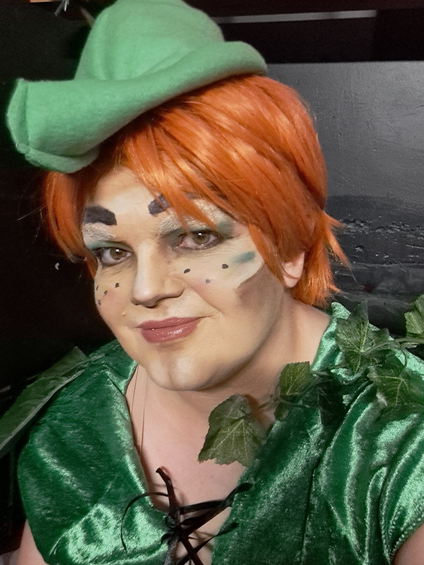 a selfie of Jamie in drag king makeup. Wearing an orange wig with a green pointy hat, green costume with fake leaves attached