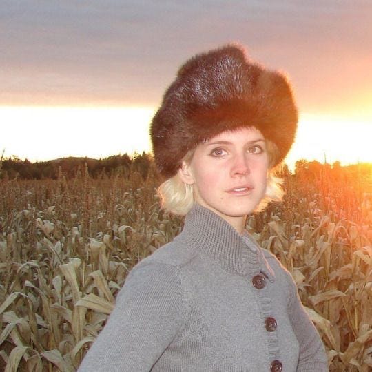 This may contain: a woman wearing a fur hat standing in a corn field at sunset with the sun behind her