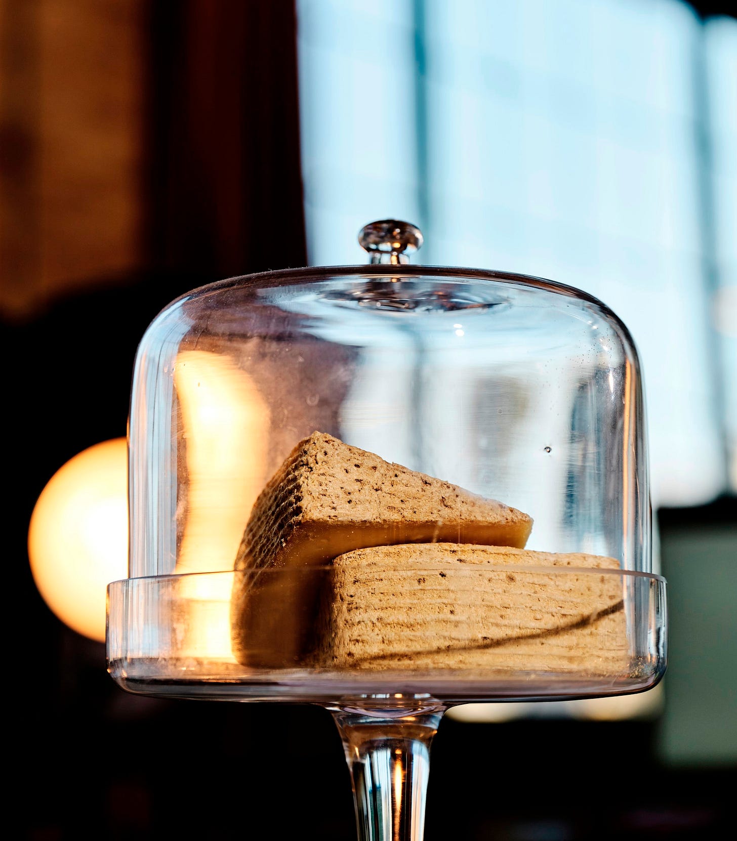 Two wedges of gruyere with a textured rind under a glass cloche on a tall pedestal, displayed in a warmly lit setting with large windows in the background.
