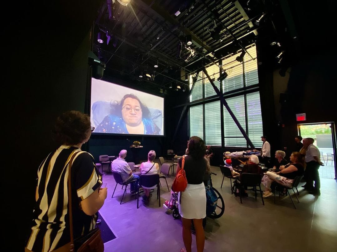 Athena's face displayed on the projection screen in a large theatre. The audience sits scattered at tables listening to her speak.