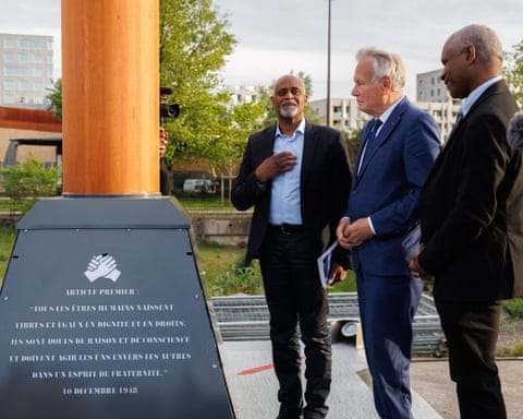 Dieudonné Boutrin, Jean-Marc Ayrault and Georges Emmanuel stand beside base of mast with clasped hand symbol and inscription beneath in French