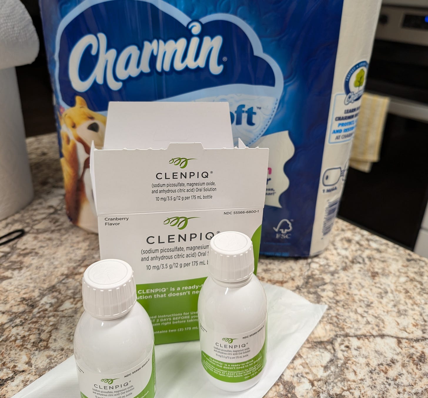  Author photo of two bottles of colonoscopy prep liquid, Clenpiq, and a big pack of Charmin toilet paper sitting on brown marbled counter top.