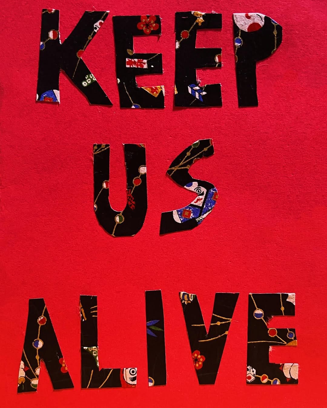 A red card with black lettering that’s says, Keep us alive. The black lettering is cut from a floral print. A red card with black lettering that’s says, Keep us alive. The black lettering is cut from a floral print.