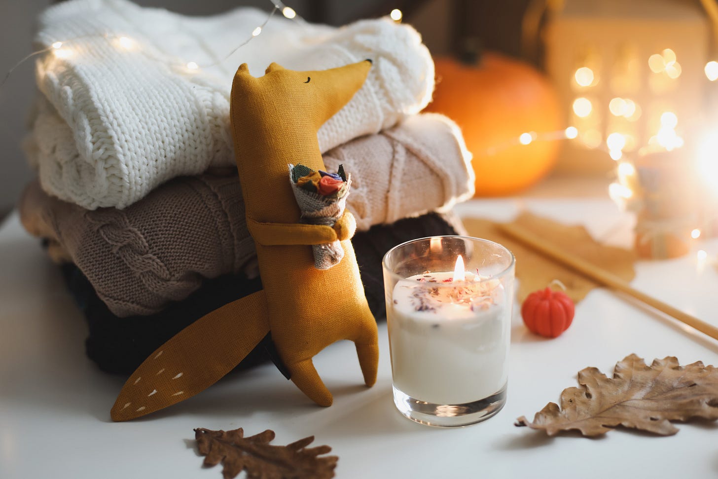 Small plush fox toy leaning against a pile of folded sweaters. the fox is holding a bouquet of flowers and there’s a lit candle in front of him. there are fairy lights over the pile of sweaters and in the background. 