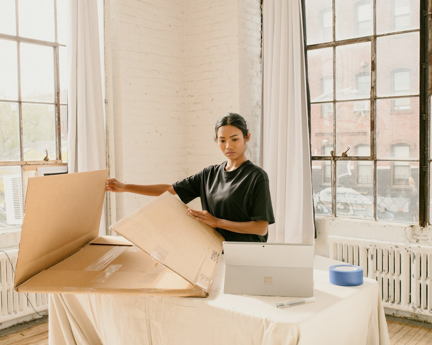 A woman in a black T-shirt folds a large cardboard box on a table next to an open laptop, in a bright room with tall windows.