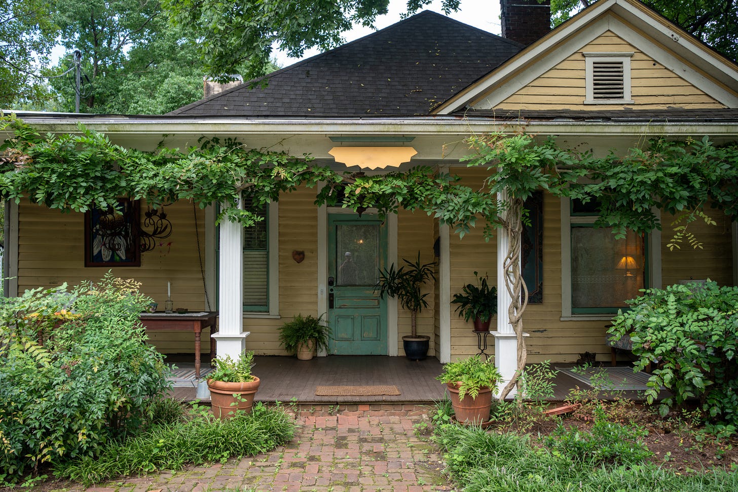 Exterior view of a yellow historic house with a covered front porch, white columns covered in greenery, and a teal front door.
