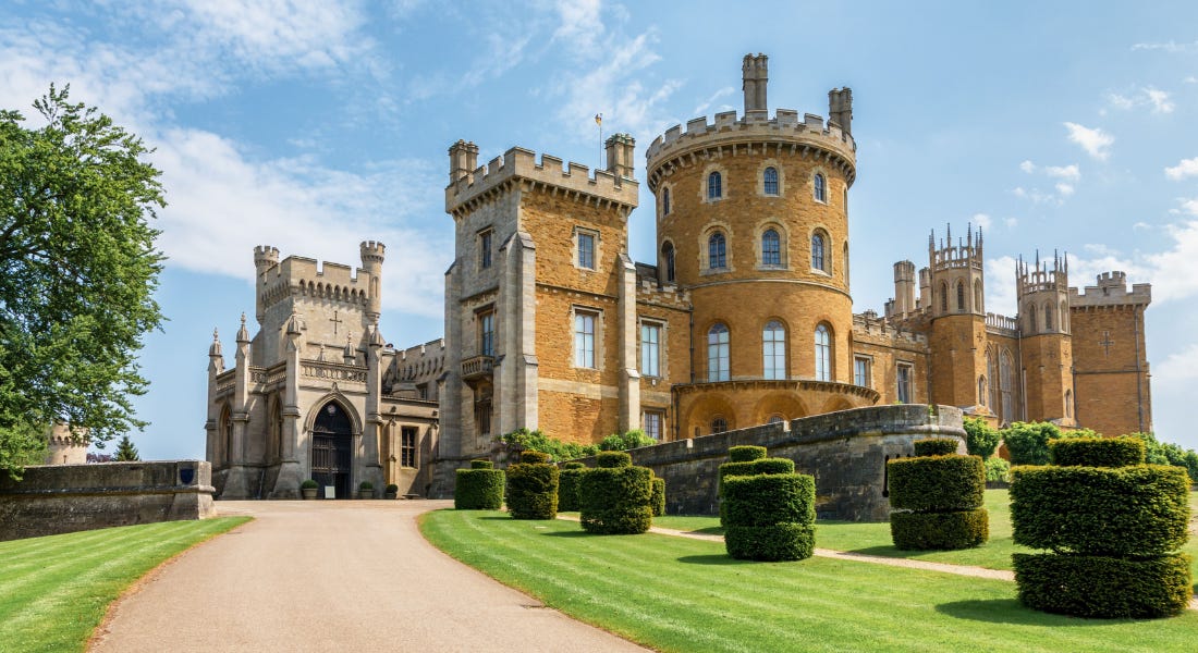 A light brown and grey castle with a driveway and lawns in front of it and a blue sky above it