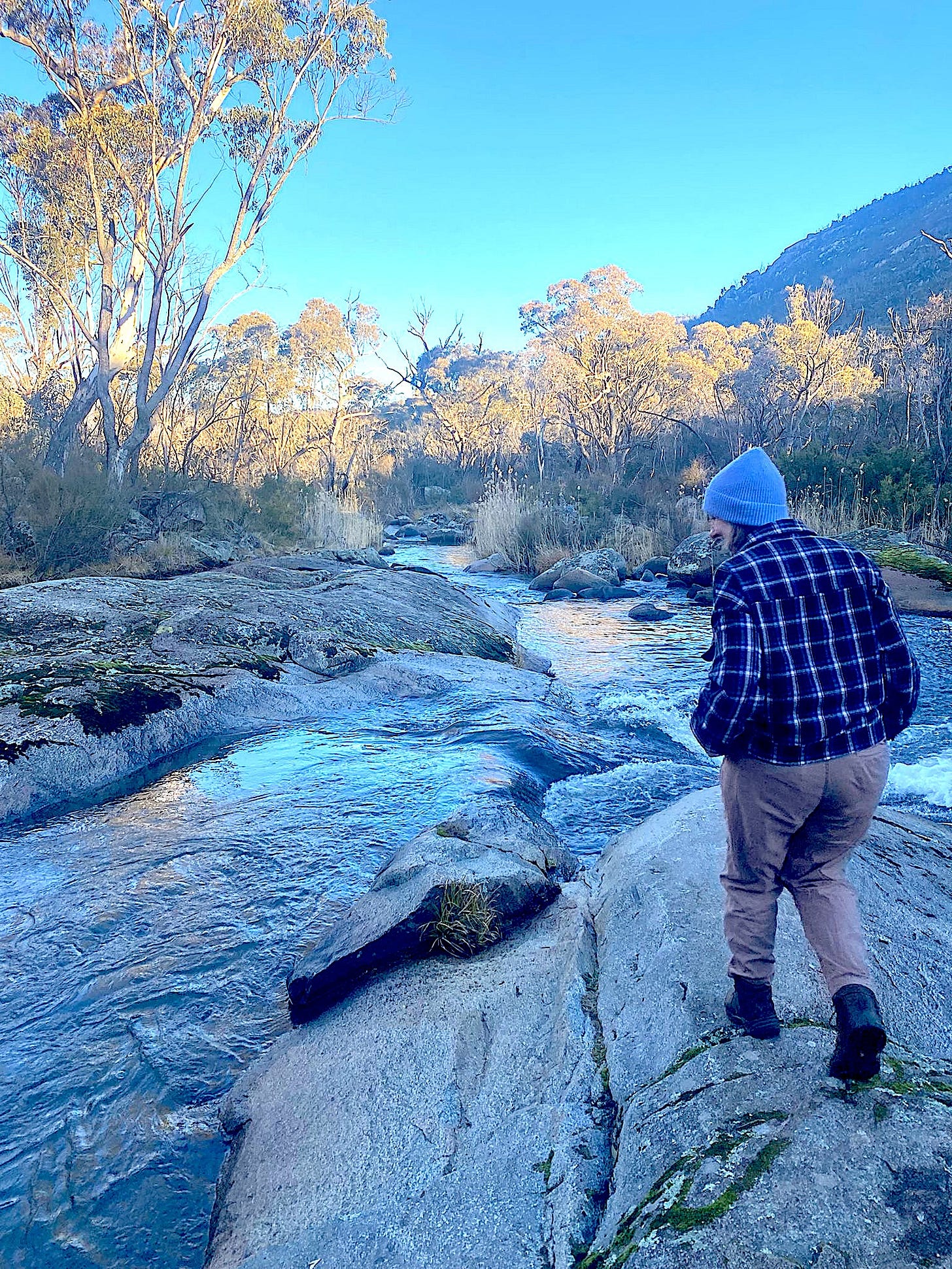 Image shows the back of a woman walking across an icy looking rockpool by a fast running creek. Around this creek is native bush and a foreboding mountain. Image shows the back of a woman walking across an icy looking rockpool by a fast running creek. Around this creek is native bush and a foreboding mountain.
