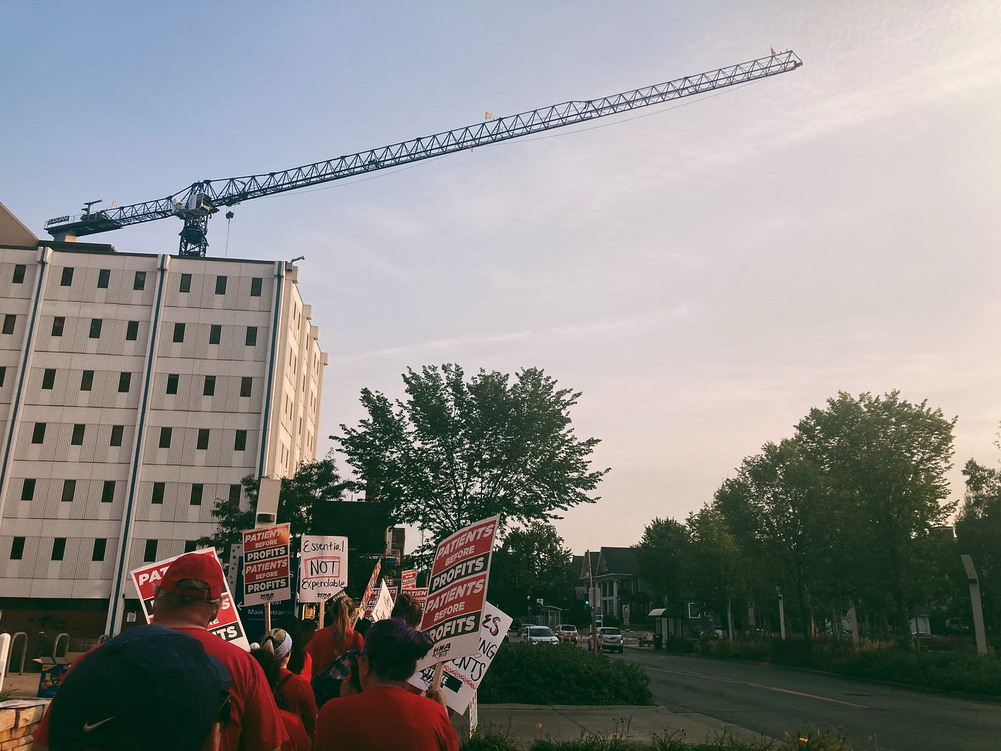 a group of people wearing red and holding picket signs that read "patients over profits" walk down the street below a construction crane and trees a group of people wearing red and holding picket signs that read "patients over profits" walk down the street below a construction crane and trees