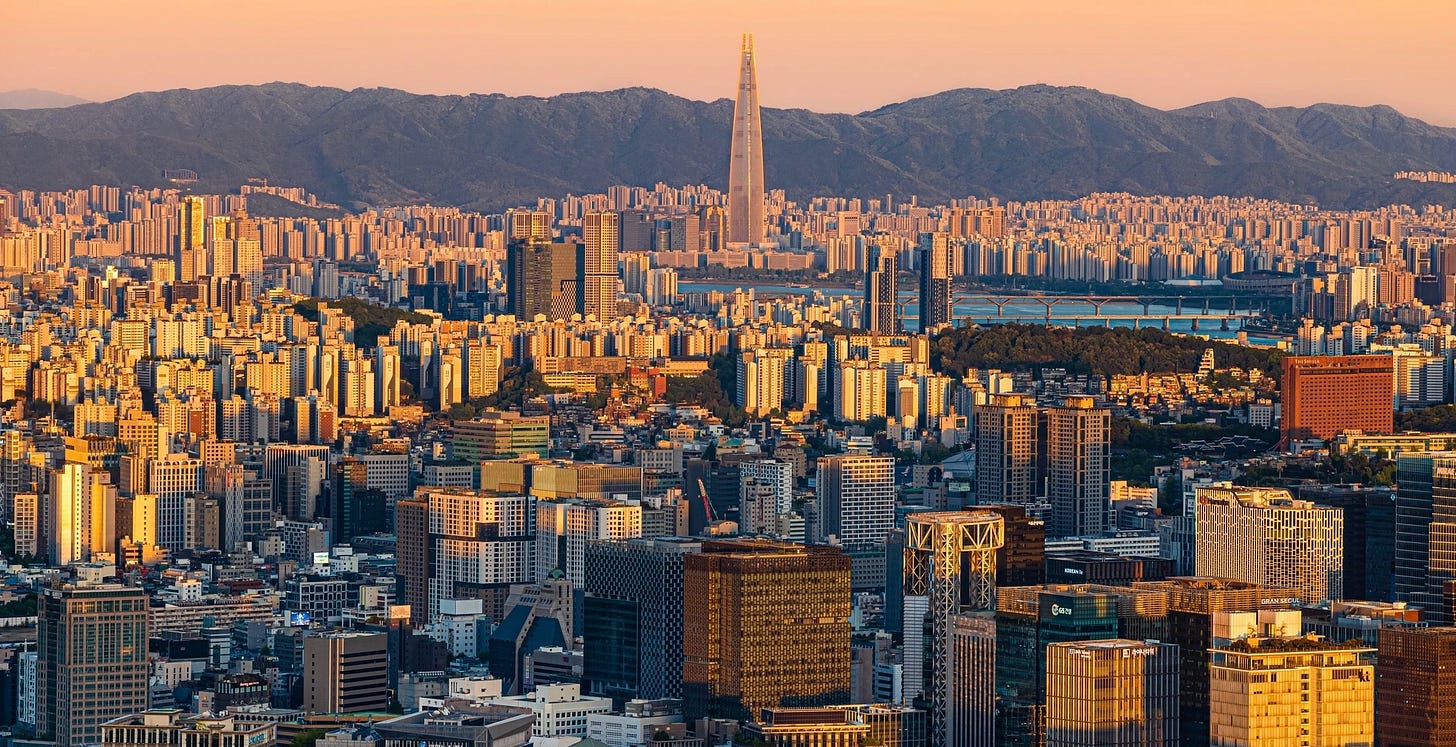 Seoul's dense skyline at golden hour, with the Han River and the Lotte World Tower rising above a sea of apartment towers, mountains behind.
