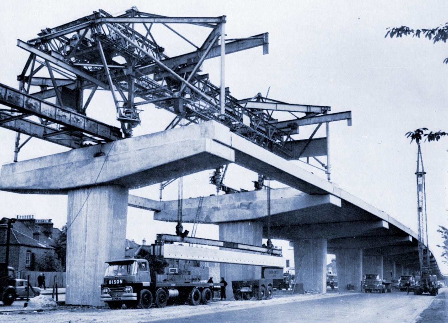 Photograph of a metal gantry structure standing atop the concrete piers of the viaduct, winching a large concrete beam from a lorry parked below.