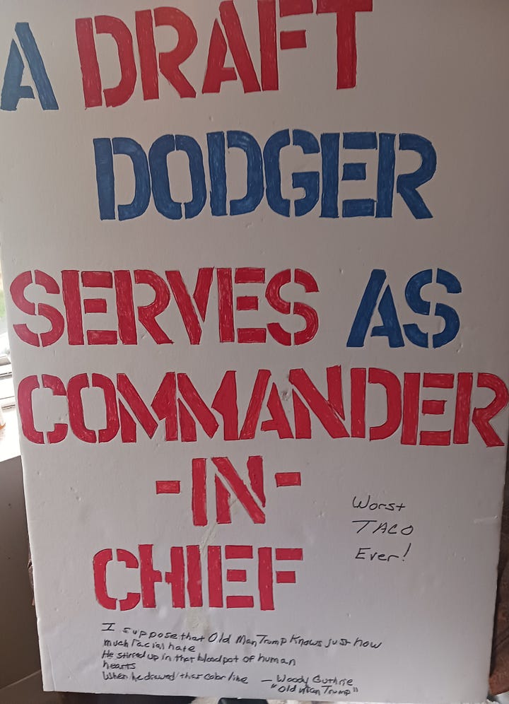 Left: Red and blue letters on white foam posterboard spell out A DRAFT DODGER SERVES AS COMMANER-IN-CHIEF. Right: Read and blue letters on white foam posterboard spell out "THIS MACHINE KILLS FASCISTS. Photos of Woody Guthrie with his guitar stating the same are next to the words THIS MACHINE and KILLS.