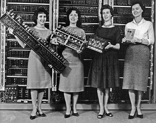 Four women stand in front of early computer equipment, each holding a piece of vintage hardware or documentation, with a backdrop of switching panels in a monochromatic photograph. Four women stand in front of early computer equipment, each holding a piece of vintage hardware or documentation, with a backdrop of switching panels in a monochromatic photograph.