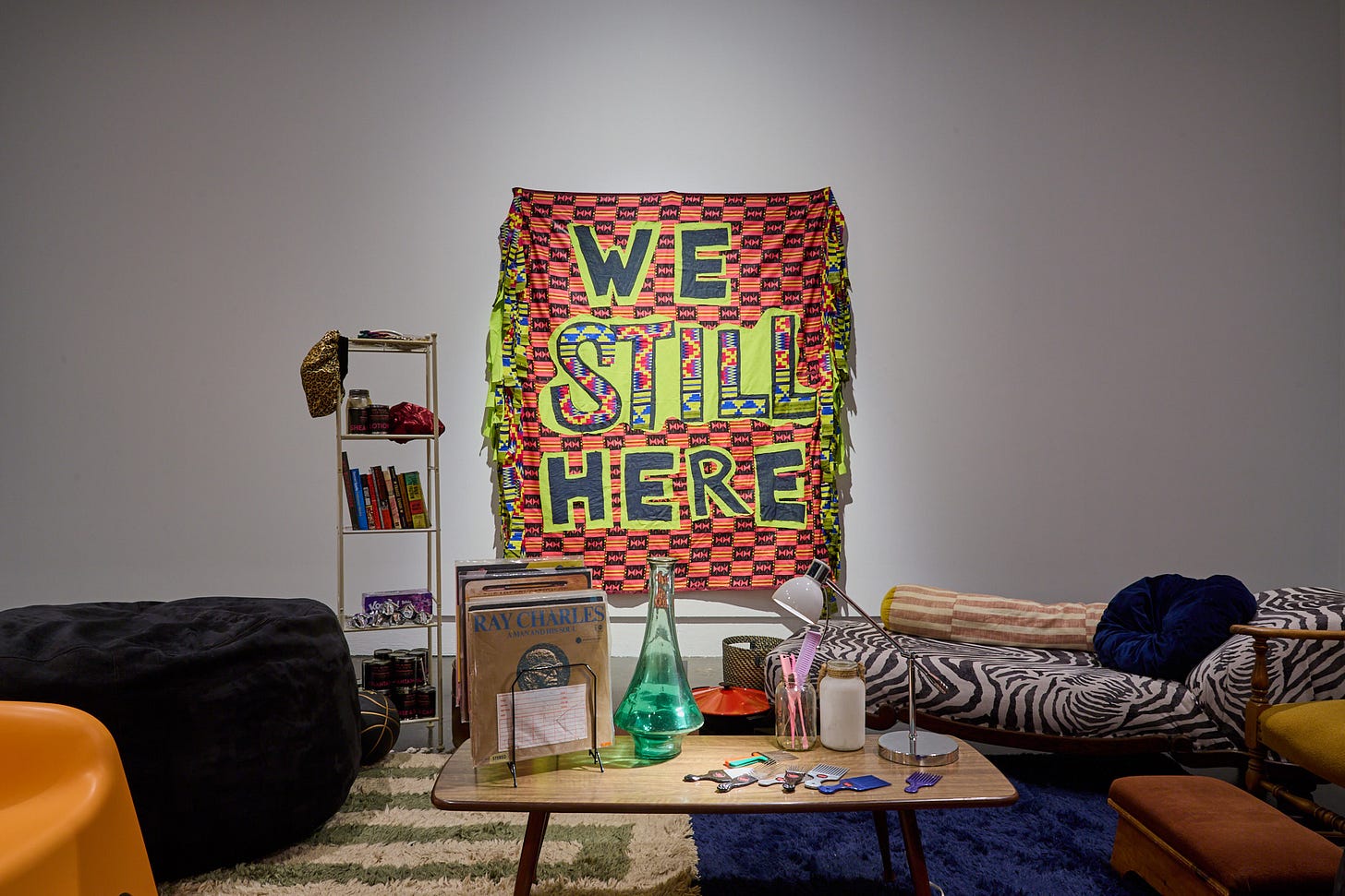 A photograph of Black Bunker by Syrus Marcus Ware. The image shows a series of chairs and sofas around a table with vinyl records by Ray Charles and a selection of afrocombs. In the background, you see a shelf featuring books and other items such as a hair bonnet and a basketball. The tapestry hanging on the wall reads '“We Still Here”.