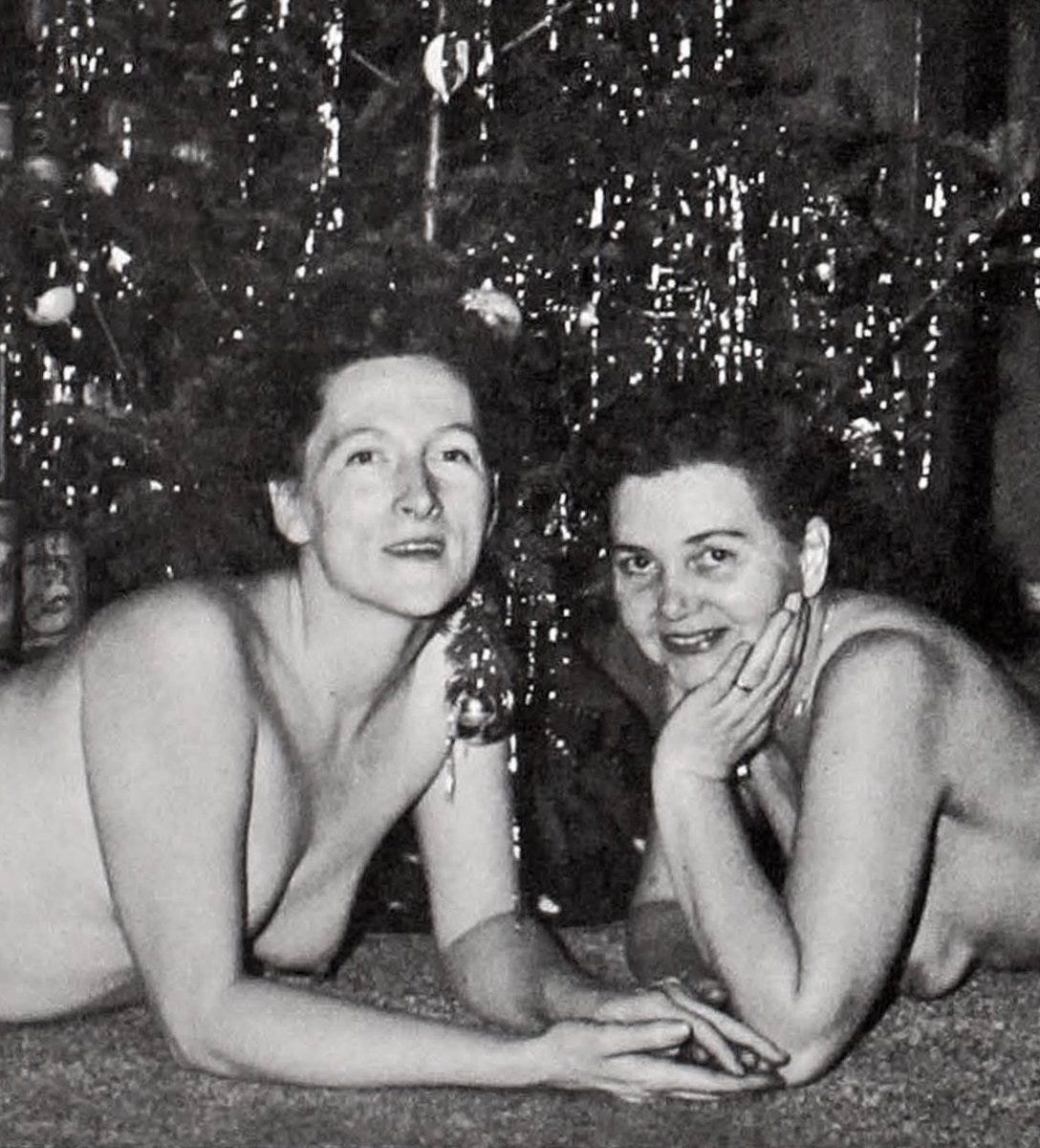 Two nude women lie on the floor in front of a decorated Christmas tree, smiling and holding hands. Tinsel and ornaments hang from the tree behind them. The photo is black and white and has a cozy, candid feel.