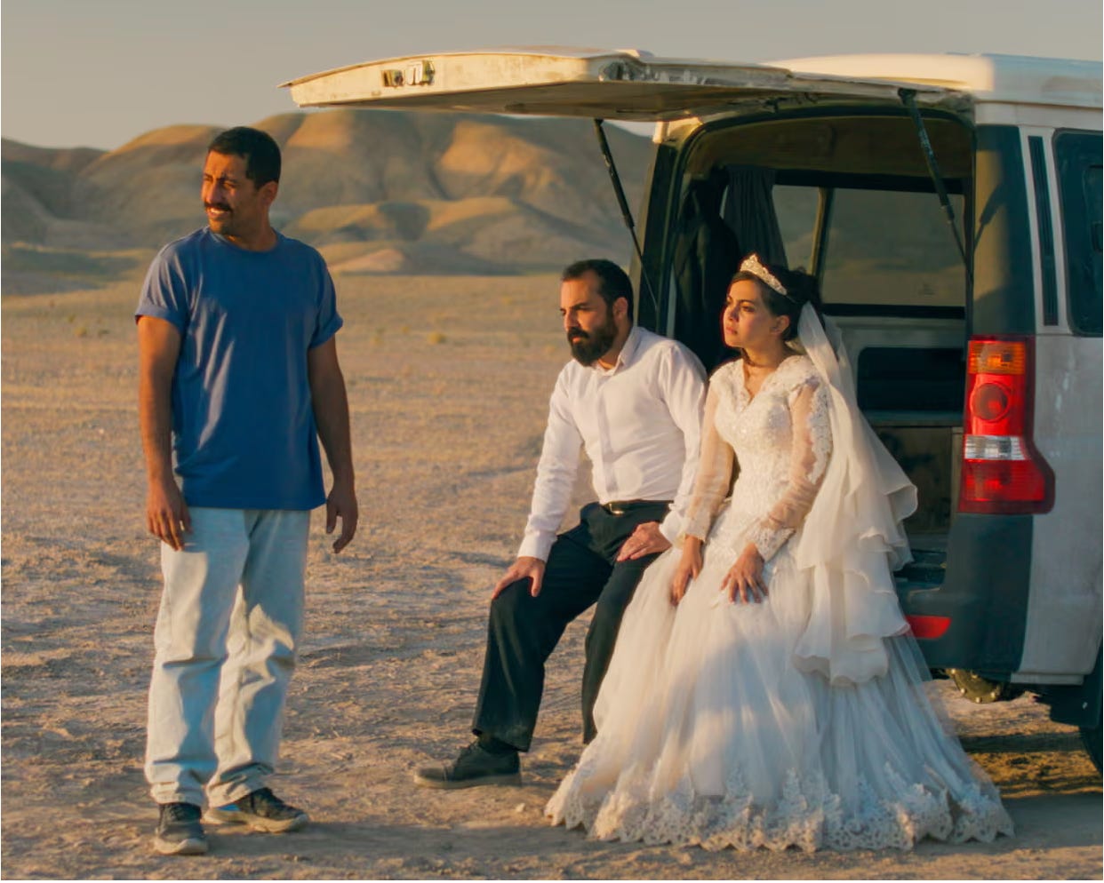 an Iranian bride and groom sit in the open hatch of a white van in the desert. an Iranian man in a blue shirt stands beside them, looking off to the side; there are mountains in the background