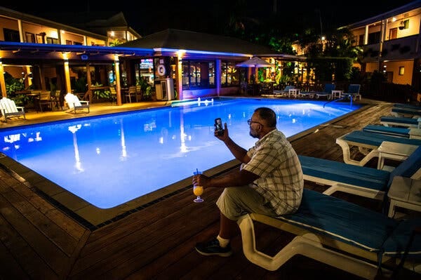 Mr. Khan sitting on a poolside lounge chair while holding his phone in one hand and a drink in the other. Mr. Khan sitting on a poolside lounge chair while holding his phone in one hand and a drink in the other.