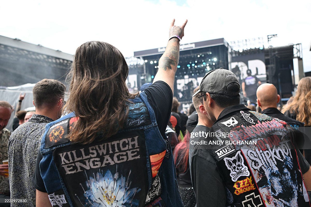 Two men in a crowd at a heavy metal concert. The stage is in the background. They are wearing cut-off denim jackets with embroidered patches sewn onto them.