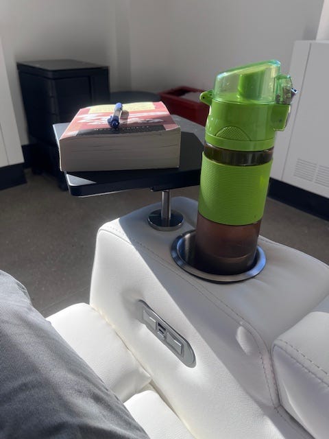 Arm of ivory upholstered recliner chair with metal cupholder, recliner buttons on the inside of the inner arm, and a black swiveling desk top. A thermos of tea sits in the cupholder; a paperback with a pen sits on the desk. Arm of ivory upholstered recliner chair with metal cupholder, recliner buttons on the inside of the inner arm, and a black swiveling desk top. A thermos of tea sits in the cupholder; a paperback with a pen sits on the desk.