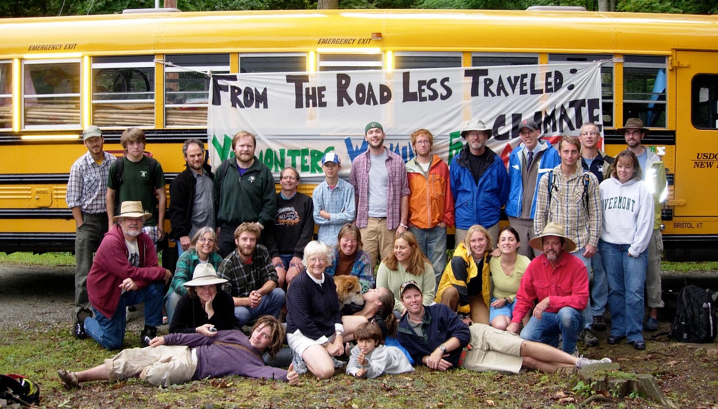 Two dozen or so people standing or sitting on grass in front of school bus with sign 'from the road less traveled'