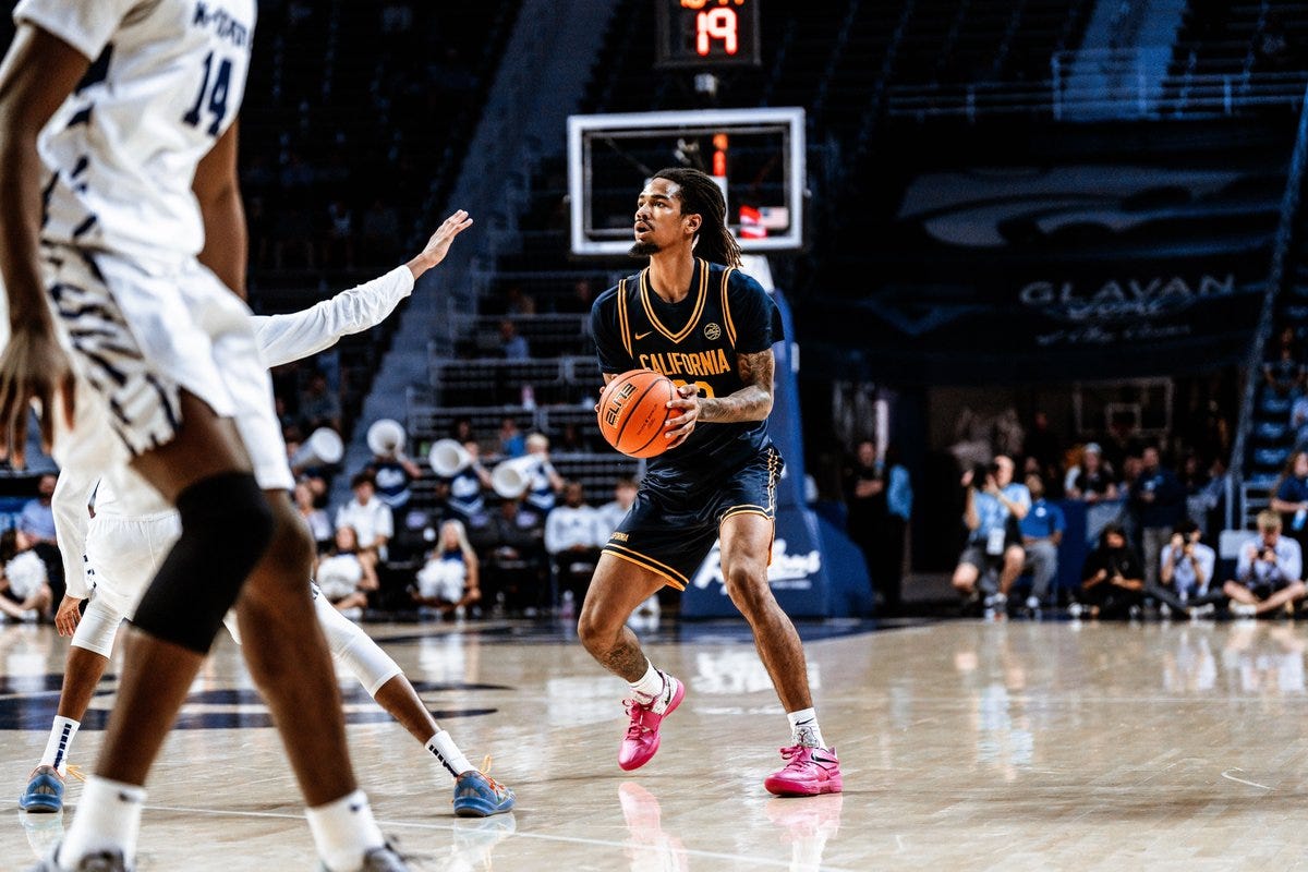 A basketball player in a navy blue California Golden Bears uniform with gold trim number 14 dribbles an orange basketball on a wooden court during a game. Teammates in similar uniforms including one with number 11 reaching out and opponents in white uniforms defend nearby. Spectators sit in blue seats in the background with a scoreboard showing 19 visible above the basket.