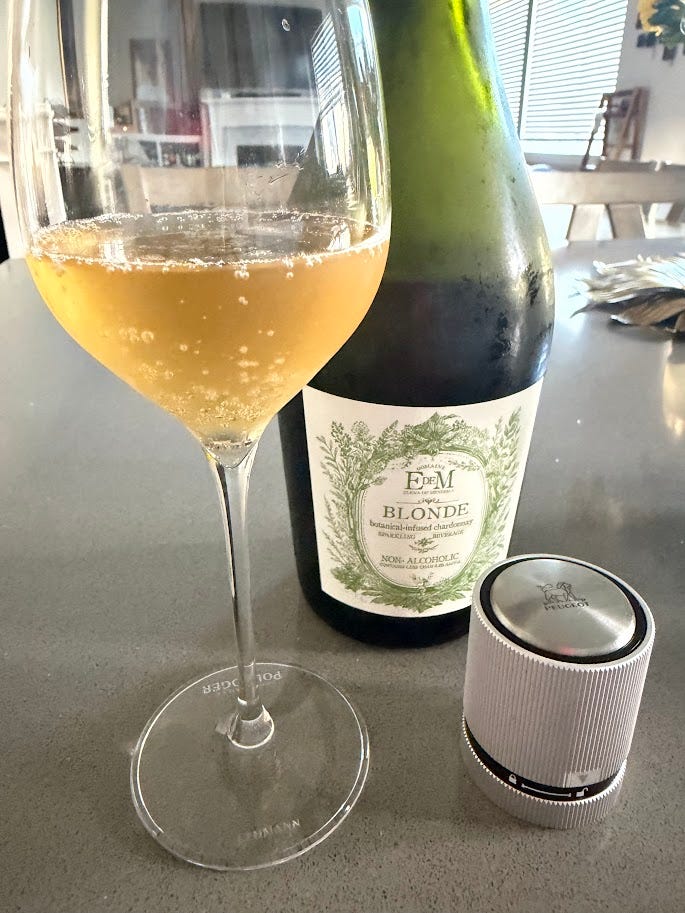 Two Domaine EdeM sparkling Chardonnays poured at home: the N.7 Uco Stones, pale gold in a stemmed glass beside its cork, and the golden-hued Blonde, non-alcoholic, poured beside a Peugeot wine preserver.