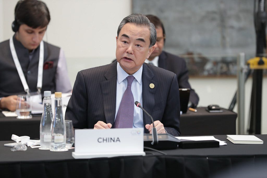 Wang Yi, ministro das Relações Exteriores da China vestindo um terno preto com risca, camisa azul clara e gravata roxa sentado em mesa em frente a placa China Wang Yi, ministro das Relações Exteriores da China vestindo um terno preto com risca, camisa azul clara e gravata roxa sentado em mesa em frente a placa China
