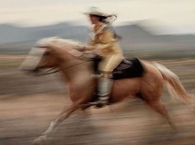 This may contain: a man riding on the back of a brown horse across a desert field with mountains in the background