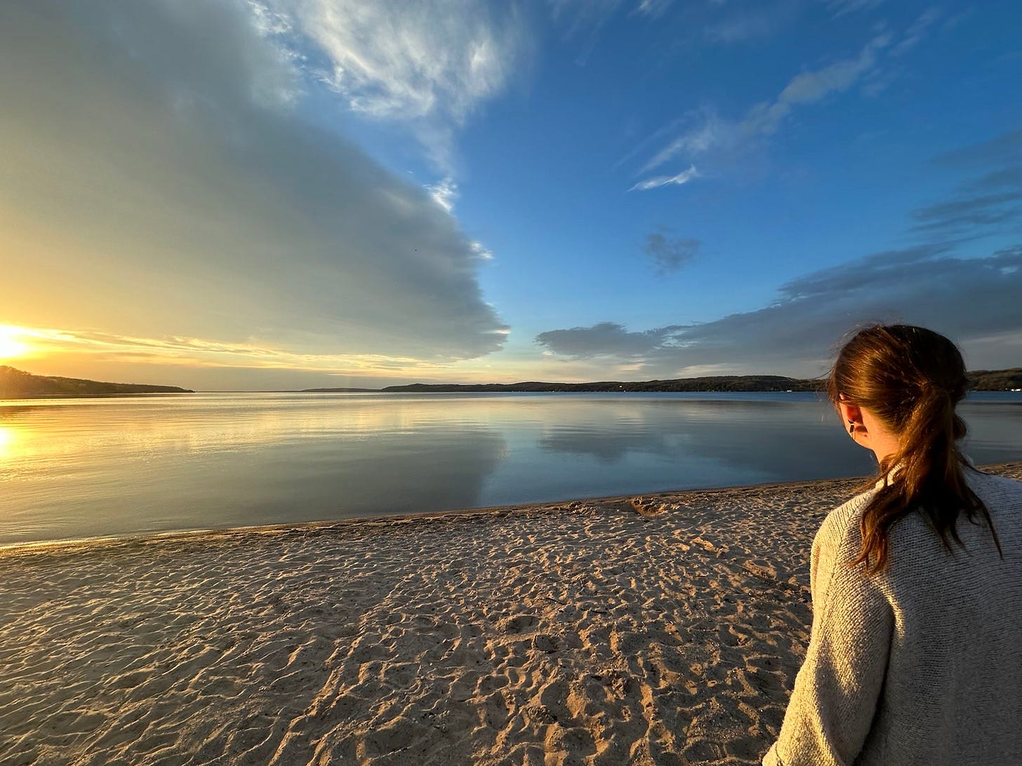 I'm standing by a lake, looking out at calm waters and the beginnings of a sunset. I'm a white woman wearing a cardigan, with a pony tail.